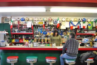 Photo couleur de l’intérieur d’un commerce avec comptoir lunch. Un homme est assis sur un tabouret, de dos, et une femme est debout derrière le comptoir.