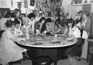 Douze femmes sont autour d’une table ronde sur laquelle quelques-unes collent des images sur un carton alors que d’autres observent et qu’une consulte une revue. 