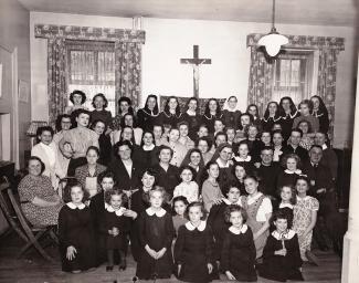 Groupe d’enfants et d’adultes en rang pour la photo dans un salon. Derrière eux, une croix avec Jésus. 