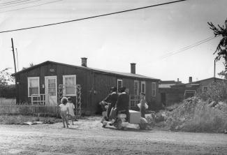 Deux femmes habillées en noir et portant un chapeau sont sur une mobylette. Elles sont devant une maison modeste entre un petit garçon et deux fillettes. 