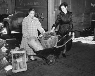 Deux femmes portant un manteau transportent des légumes avec une brouette.