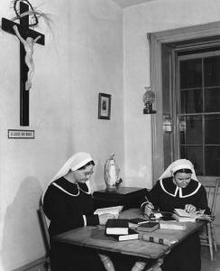 Deux femmes attablées, portant costume et voile blanc, consultent des livres et prennent des notes. Sur le mur, une grande croix avec Jésus. 