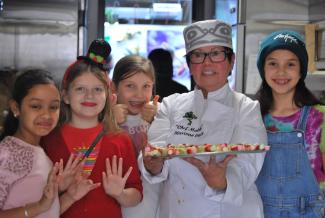 Une chef tenant un plateau de nourriture est entourée de quatre petites filles souriantes.