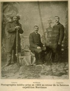 Photographie sépia de trois hommes en vêtements civils dans un décor de studio photographique. L’homme de gauche porte un chapeau et s’appuie sur un fusil.