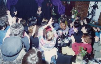 Photo couleur d’un spectacle à l’intérieur d’un bar. Une chanteuse se trouve à l’avant, et devant elle, des femmes assises à des tables applaudissent.