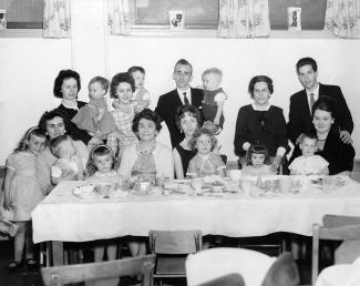 Sept femmes sont assises ou debout devant une table avec nappe et couverts. Elles sont accompagnées de deux hommes et de neuf enfants. 