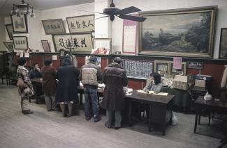 Photo couleur montrant une dizaine de personnes d’origine chinoise dans une grande salle, certaines sont debout, de dos, devant une table, d’autres assises derrière la table. 