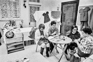 Quatre femmes sont assises et tricotent. Une table se trouve entre elles. On voit plusieurs de leurs réalisations dans la pièce derrière elles.