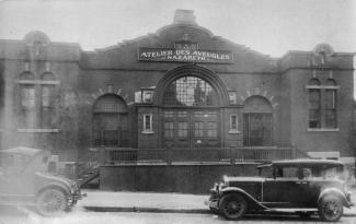 Grand immeuble d’un seul étage, avec arches vitrées au-dessus des portes. Des voitures anciennes sont stationnées devant