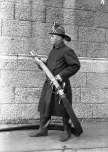 Photographie en pied d'un pompier en uniforme tenant un boyau et une lance d'incendie.