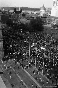 Foule amassée dans la rue entre deux places publiques. Trois drapeaux flottent au vent. À l’arrière-plan, on reconnaît le Château Ramezay, la chapelle Notre-Dame-de-Bon-Secours et le marché Bonsecours. 
