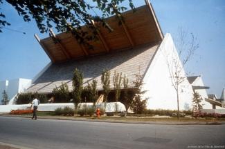 Photographie couleur montrant un pavillon de forme pyramidale dont une excroissance du toit s’élance vers le ciel. Des arbres et des éléments de terrassement sont présents devant le pavillon à l’avant-plan.