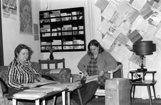 Deux femmes sont assises devant une table basse. Autour d’elles, on voit des affiches sur la contraception et sur l’Année internationale de la femme.