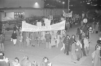 Des dizaines de personnes marchent dans la rue. Des manifestants tiennent une affiche pour le regroupement des garderies sans but lucratif.