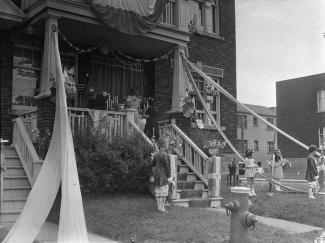 Deux garçons et deux fillettes se tiennent au bas du balcon d'une résidence décorée pour la Fête-Dieu. Sur le balcon, deux femmes déposent des fleurs sur un tabernacle.