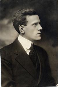 Photographie en noir et blanc montrant un homme de profil. Il porte un complet et une cravate noirs avec une chemise blanche à haut col. 