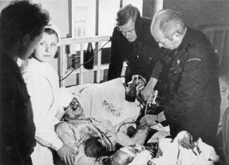 Photo en noir et blanc de deux médecins faisant une transfusion sanguine à un blessé, et une infirmière et un homme sont de l'autre côté du lit.