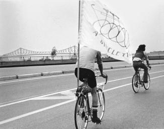 Deux cyclistes circulent avec le pont Jacques-Cartier en arrière-plan, l'un porte un drapeau du Monde à bicyclette.