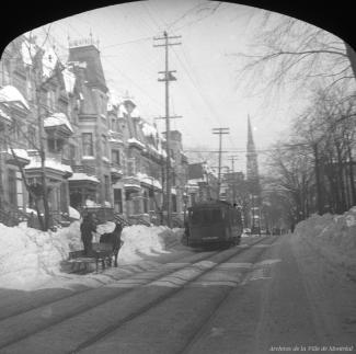 Photographie d'un tramway sur la rue Saint-Denis à Montréal, l'hiver. On y aperçoit les maisons longeant le côté est de la rue Saint-Denis, le clocher de l'église Saint-Jacques, ainsi qu'un homme monté sur un petit traîneau tiré par un cheval.