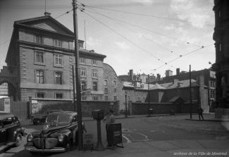 Photographie du séminaire Saint-Sulpice situé sur la rue Notre-Dame à Montréal. On y voit des voitures taxi stationnées en bordure de la place d'Armes, ainsi que des gens près des voitures. On remarque aussi les câbles servant à la circulation des tramway