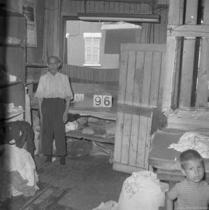 Photo en noir et blanc de l’intérieur d’une buanderie chinoise. Un homme âgé d’origine chinoise est debout au fond de la pièce. On voit un enfant, en partie coupé, dans le coin en bas à droite.