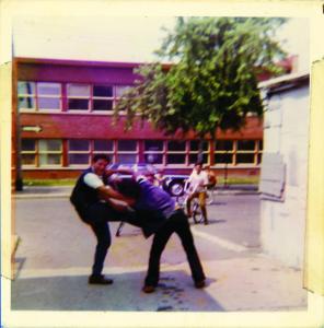 Photo couleur de deux garçons se chamaillant dans une ruelle, des garçons derrière eux sur leur vélo, et une école à deux étages en arrière plan.