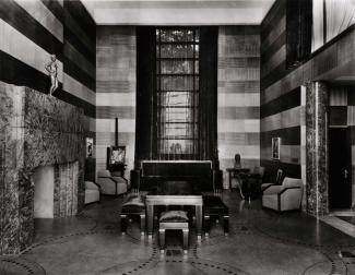 Photographie en noir et blanc montrant l’intérieur d’un salon luxueux. Les murs sont de deux teintes différentes, l’une pâle et l’autre foncée. Il y a un foyer à gauche dont le revêtement est de marbre. Il y a des fauteuils disposés sur les pourtours.
