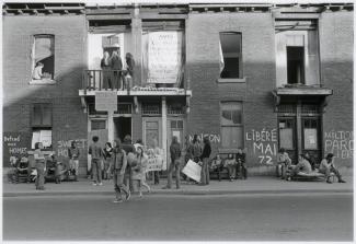 Photo en noir et blanc montrant une manifestation devant des bâtiments en briques. Plusieurs personnes sont rassemblées et tiennent des pancartes. Des banderoles et des inscriptions sont visibles sur les murs extérieurs des édifices.