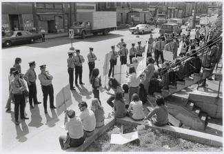 Photographie en noir et blanc montrant un groupe de personnes assises et debout sur le trottoir devant un bâtiment. Plusieurs portent des pancartes et de nombreux policiers se tiennent devant eux.