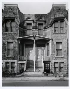 Photo en noir et blanc montrant la façade de deux résidences jumelées. La toiture est ouvragée et les portes avant sont surplombées d’un petit toit en bois.