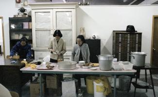 Trois femmes d’origine chinoise sont debout derrière des tables dans une grande salle à préparer de la nourriture.