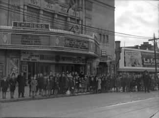 Cette photographie, prise le 30 octobre 1943, montre une file d’enfants devant l’Empress Theatre. Les enseignes lumineuses incitent à soutenir l’effort de guerre canadien par l’achat de bons de guerre. 