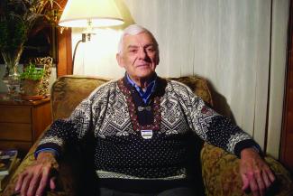 Photographie couleur d’un homme âgé aux cheveux blancs assis dans un fauteuil.