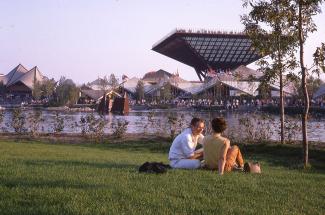 Photo couleur montrant le pavillon du Canada en arrière-plan. En avant-plan, un jeune couple est assis dans l'herbe. 
