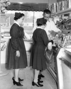 Deux femmes portant un costume noir et un chapeau assorti, dans un commerce. Un employé montre un plat de légumes qu’une des deux femmes observe.
