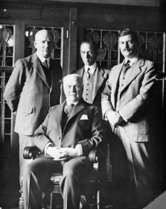 Photo en noir et blanc de quatre hommes en complet, trois sont debout et un est assis devant.