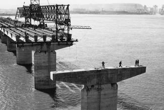 Construction d’un pont au milieu d’un cours d’eau, avec la ville en arrière-plan. En avant-plan, on aperçoit quatre hommes sur une des piles qui n’est pas encore reliée aux autres sections. 