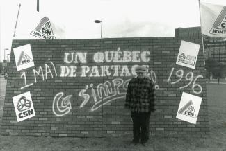 Un homme pose devant une murale où on peut lire le slogan « Un Québec de partage, ça s’impose. 1 mai 1996 ».