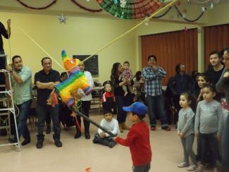 Des adultes et des enfants dans une salle communautaire observent un enfant tapant sur une piñata.