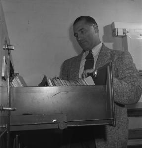 Photographie en noir et blanc d’un homme se tenant debout derrière un classeur. 