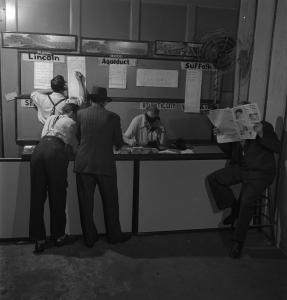 Intérieur d’une maison de paris : deux hommes sont derrière un comptoir, un debout et l’autre assis au téléphone, et trois hommes sont devant, deux debout et un assis avec un journal ouvert devant son visage. 