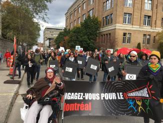 Des dizaines de personnes marchent dans la rue tenant des affiches. On peut lire sur certaines d’entre elles : « Engagez-vous pour le communautaire ».