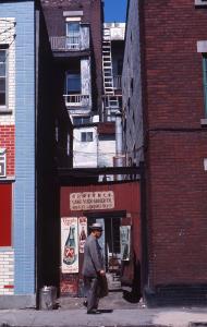 Un homme passe devant une toute petite épicerie prise entre deux édifices plus hauts. 