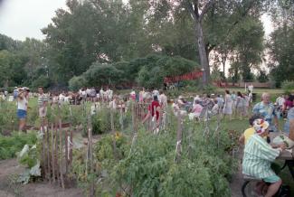 Plusieurs dizaines de personnes sont rassemblées à côté des parcelles d’un jardin. Certaines sont attablées à une table à pique-nique, d’autres sont assises sur des chaises pliantes.