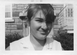 Photo en noir et blanc en gros plan d’une jeune fille souriante. Derrière elle, un immeuble à appartements.  