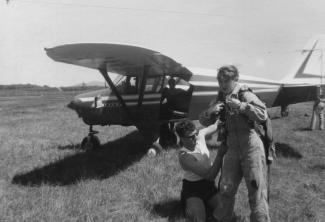 Photo noir et blanc montrant un petit avion dans un champ, une jeune fille qui met l’équipement pour faire du parachute et une personne qui l’aide.