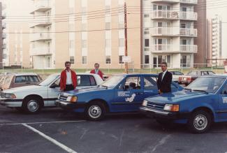 Photo couleur prise dans un stationnement montrant trois hommes prenant la pose avec trois véhicules de la Communauté urbaine de Montréal. Un édifice à logements se trouve à l’arrière-plan.