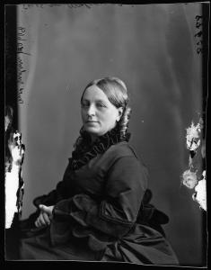 Portrait en noir et blanc d’une femme assise légèrement de profil. Elle porte une robe noire et est assise devant un fond uni. 