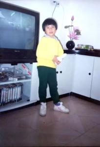 Family photo of a child standing in a living room, with a television and wall cabinet behind him. 