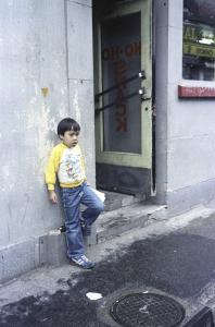 Un jeune enfant d’origine chinoise se tient debout contre le mur à côté de la porte d’entrée d’un restaurant. On peut lire Ho Ho Snack à l’envers sur la porte vitrée.